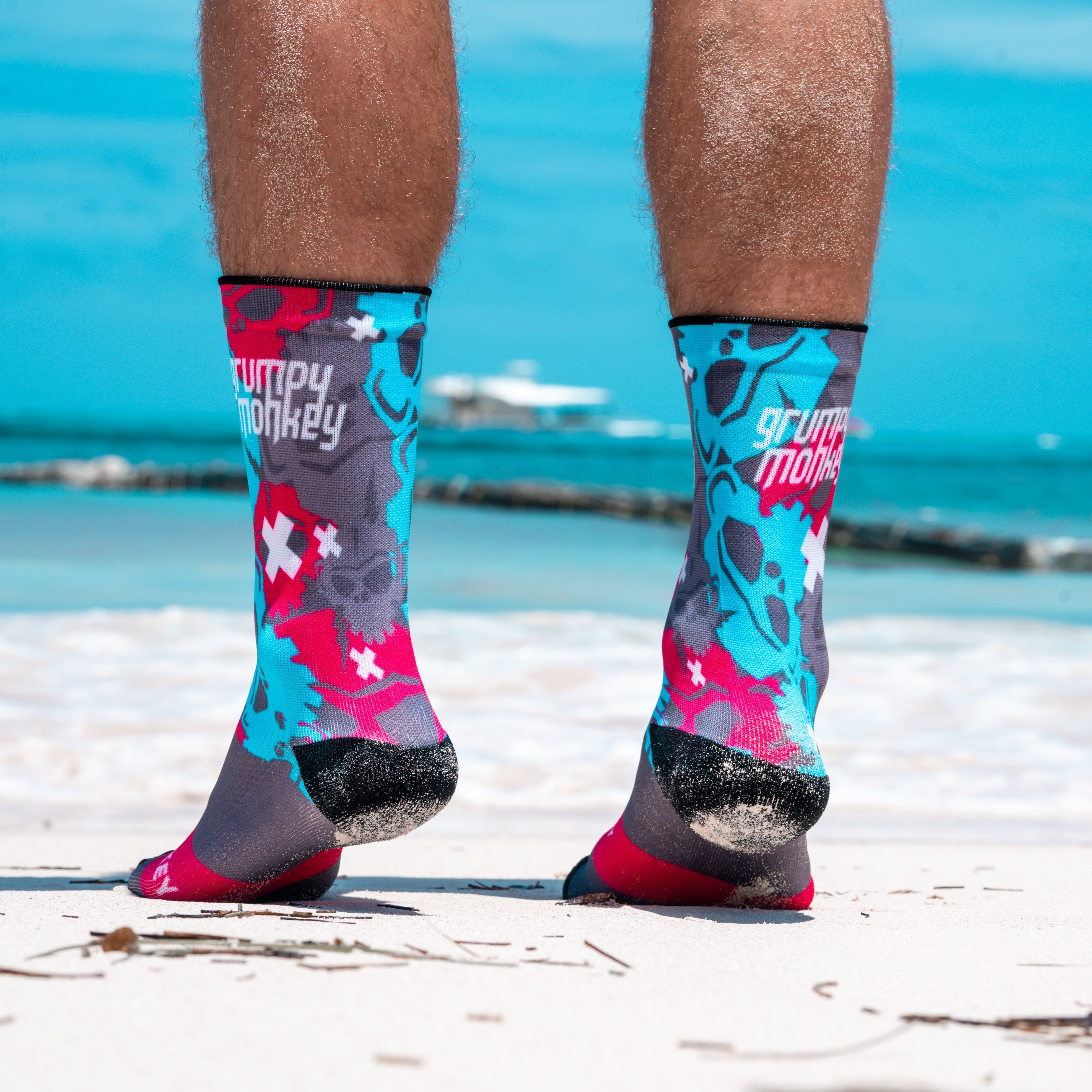 This photo shows a man with strong claves standing on his toes wearing Grumpy Monkey Funky Dab socks on the beach sand. The socks design has various monkey skulls designed thoughout the socks with red, light blue and grey skulls and white crosses in various sizes. The photo also showcases the word Grumpy Monkey in white on the side top calf. The raised heels shows the black heel supports from behind on a lovely Punta Cana beach in the Dominican Republic.